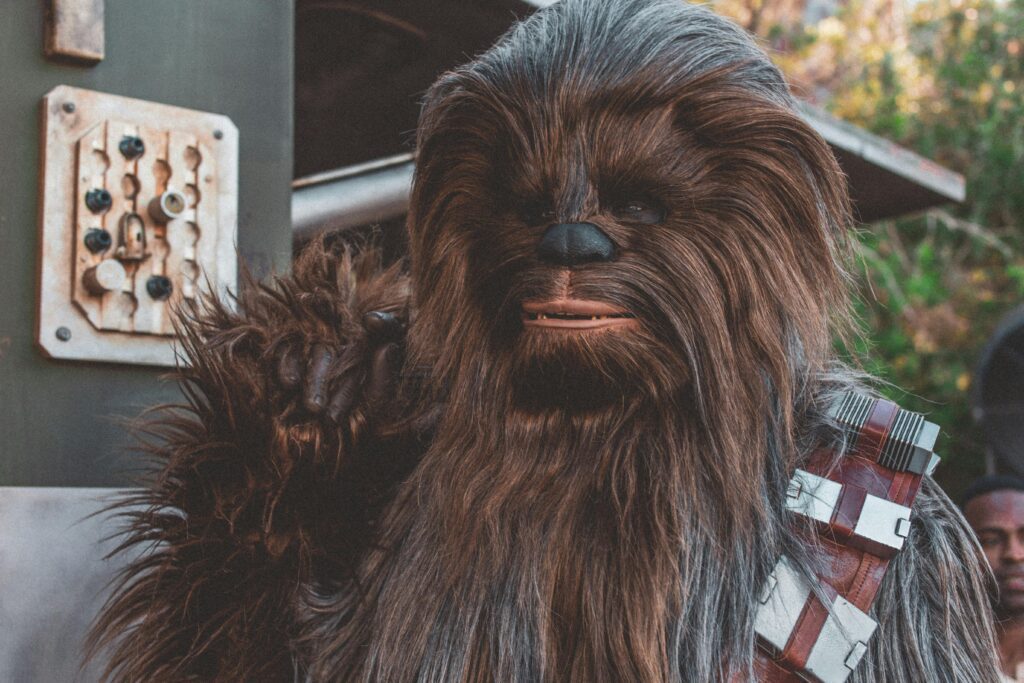 A character dressed as Chewbacca from Star Wars, featuring brown fur and a bandolier across the chest, stands outside near a futuristic-themed building. The background includes greenery and electronic panels.