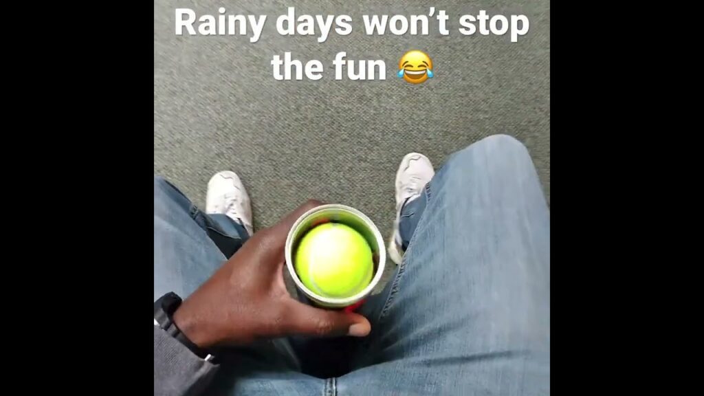 A person sits indoors holding a container with a tennis ball, wearing jeans and sneakers, with text above saying, "Rainy days won’t stop the fun 😂".