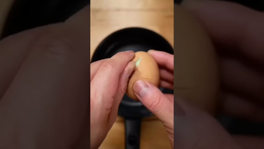 Hands cracking an egg with a small sticker over a frying pan on a wooden surface.