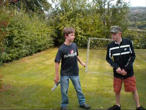 Two young people are standing in a grassy yard with a soccer goal in the background. One is holding a sword prop and wearing a dark shirt and jeans, while the other is in a black jacket, camouflage cap, and red shorts. Trees line the edge of the yard.