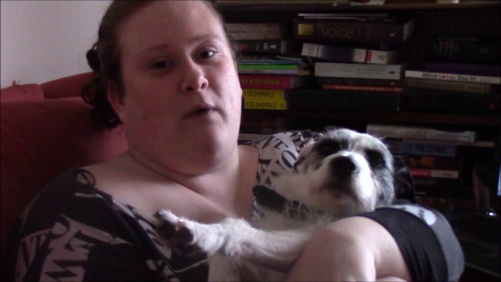 A person with light skin holds a small, black and white dog in their arms while sitting on a red couch. Behind them is a stack of books on a shelf.