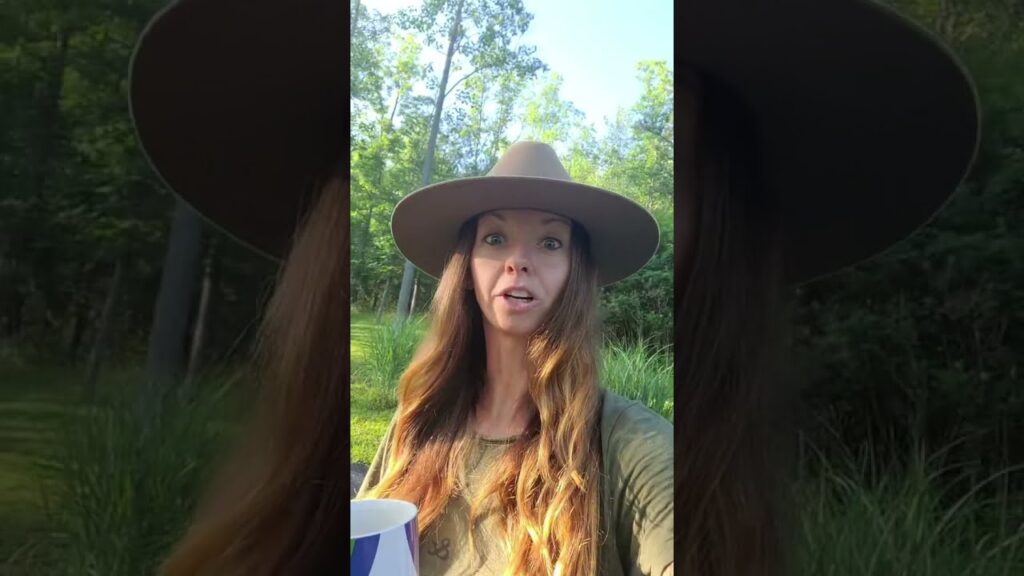 A person with long hair wearing a wide-brimmed hat and holding a blue and white mug stands outdoors. They are surrounded by greenery and tall trees, under a clear sky.