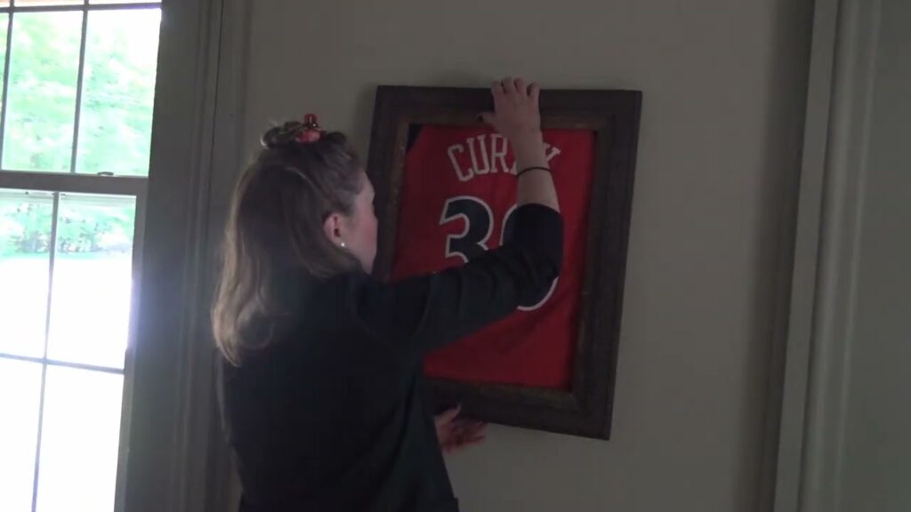 A person is hanging a framed sports jersey with the number 30 and the name "Curry" on a wall next to a window. The room is softly lit by natural light coming through the window.