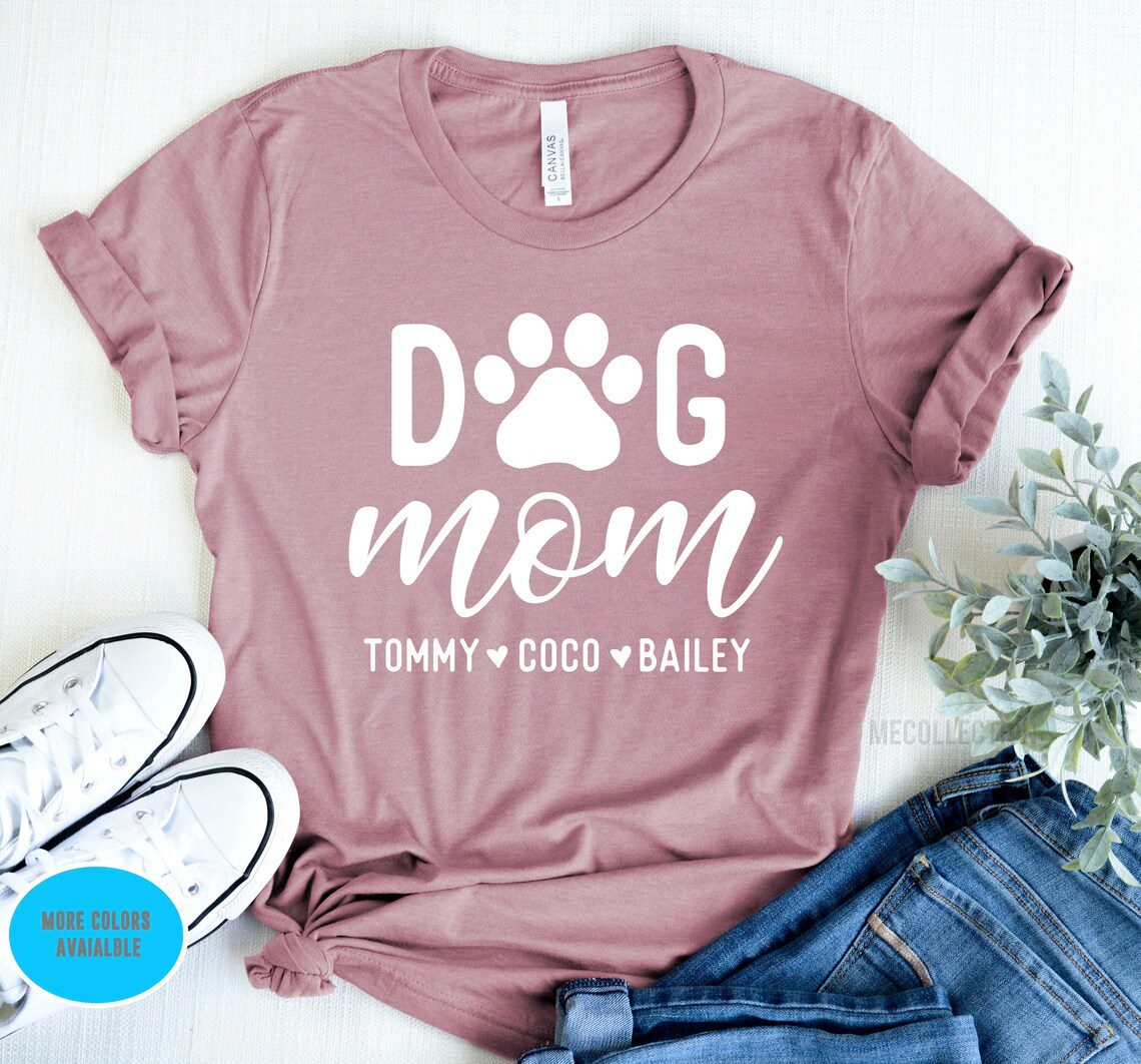 The Personalized Names Dog Mom Shirt in mauve features "Dog Mom" in white letters, with a paw print replacing the "O," followed by the names Tommy, Coco, and Bailey. It's styled with white sneakers and jeans against a light fabric background.