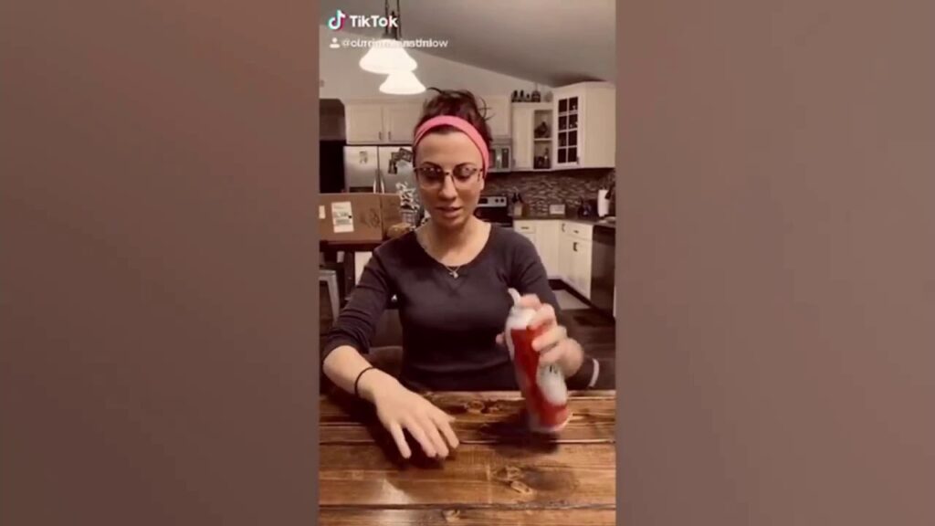 A person with a pink headband sits at a wooden table in a kitchen, holding a can of whipped cream. There are kitchen cabinets and appliances in the background.