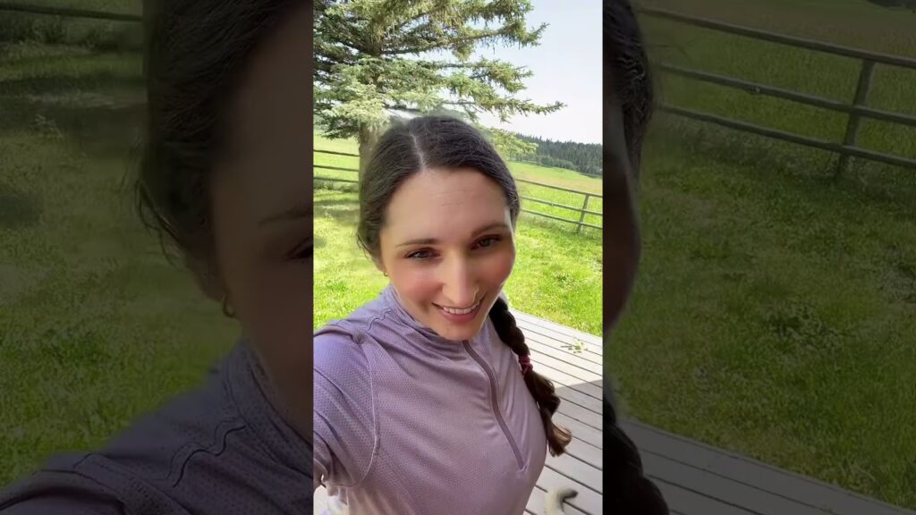 A person with long hair in a braid, wearing a purple top, smiles at the camera. They are standing outside on a wooden deck with a grassy field and fence in the background. A tree is visible on the left.