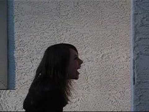 A person with long dark hair appears to be shouting or expressing intense emotion while facing a textured, light-colored wall.