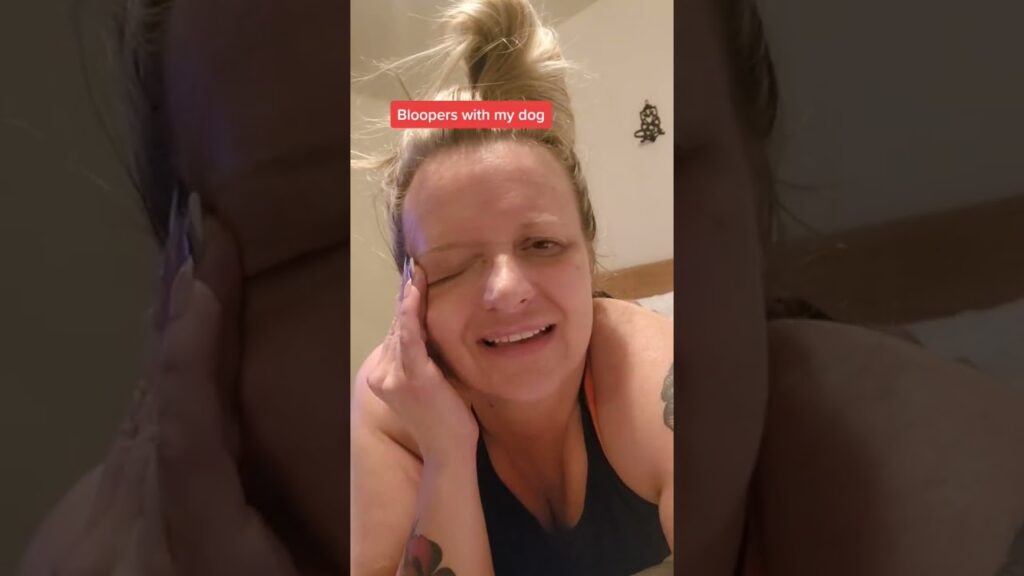 A person with a top bun hairstyle smiles while squinting one eye. They are wearing a tank top and resting their face on their hand. Text on the image reads "Bloopers with my dog." Tattoos are visible on their arm.