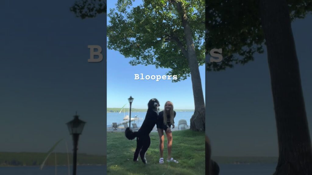 A person in a dog costume jumps next to a smiling woman standing by a large tree, with a lake and boats in the background. The word "Bloopers" is overlaid at the top.