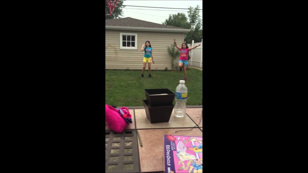 Two children play in a backyard, one wearing a blue shirt with yellow shorts, and the other in a pink shirt with red shorts. In the foreground, a table holds a water bottle, plant pots, and a book.