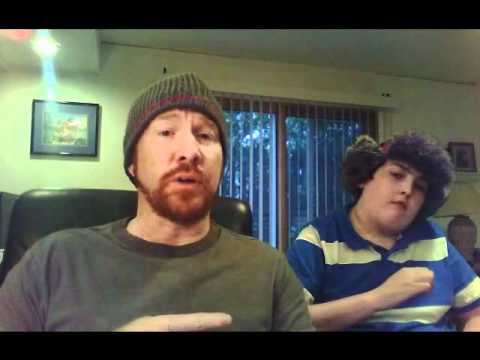 A bearded man wearing a beanie and a boy in a striped shirt, both indoors. The man gestures with his hand while speaking. Behind them is a window with blinds and a framed picture on the wall.