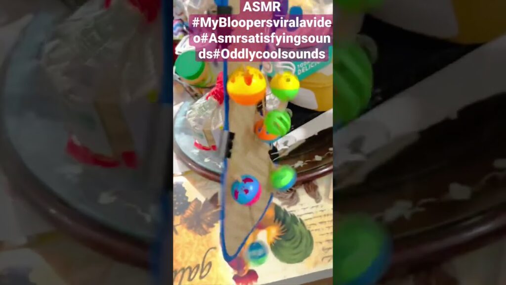 A colorful toy with small balls on a winding path is captured in motion, creating satisfying sounds. Text overlays mention "ASMR" and various hashtags like #MyBloopersviralavideo and #Asmrsatisfyingsounds.