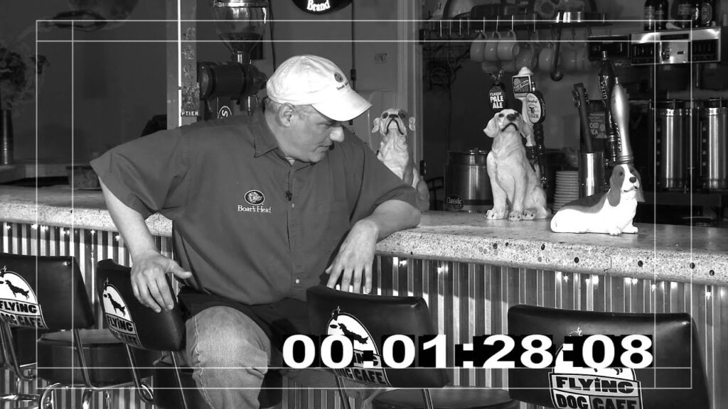 A man in a cap and uniform leans on a bar counter at Flying Dog Cafe, facing two dog statues. The scene is in black and white, with a timer in the bottom right corner displaying "00:01:28:08.