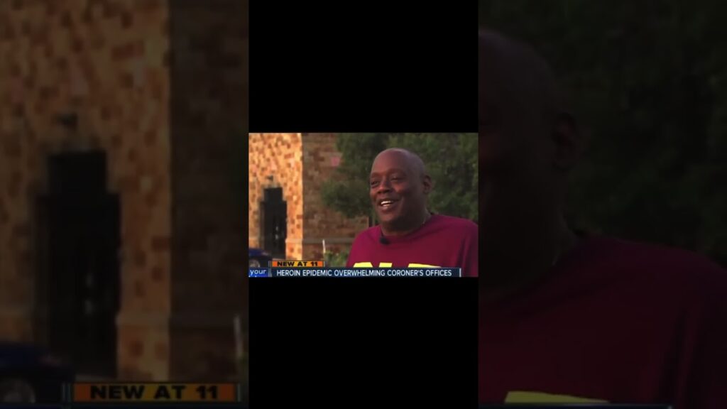 A person in a maroon shirt stands outdoors smiling. A news ticker at the bottom reads, "HEROIN EPIDEMIC OVERWHELMING CORONERS OFFICES." Stone buildings and greenery are in the blurred background.