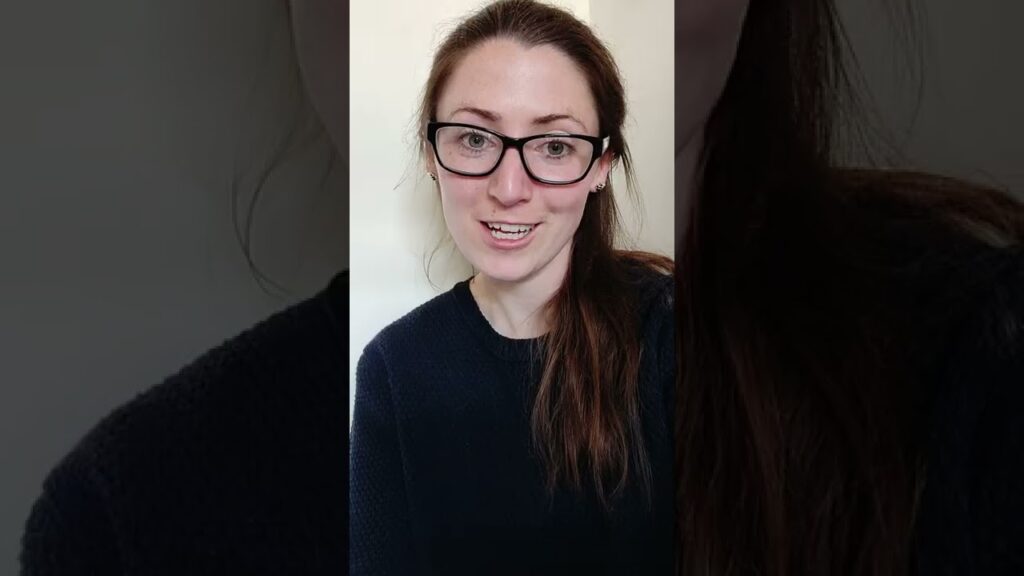 A woman with long brown hair wearing glasses and a black sweater smiles at the camera. She is indoors with a neutral background.