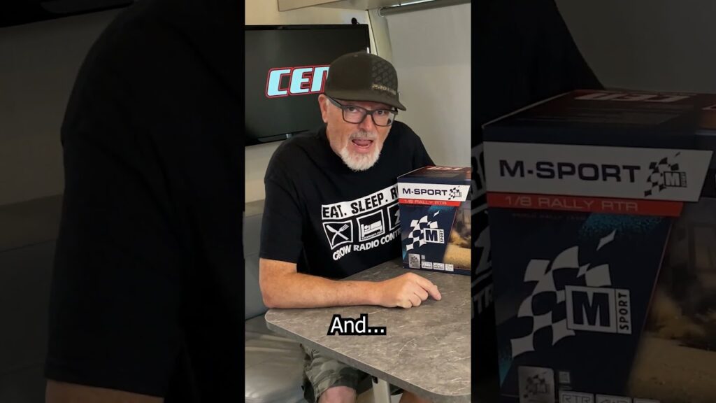 A man with a beard and glasses, wearing a cap and a black T-shirt, is sitting indoors at a table. He is smiling and holding a box labeled "M-Sport 1/8 Rally RTR." A TV screen with the word "CEN" is in the background.