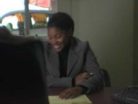 A person in a gray blazer sits at a table, smiling and holding a notebook. There is a window in the background, partially showing outside. A computer keyboard is visible in the foreground.