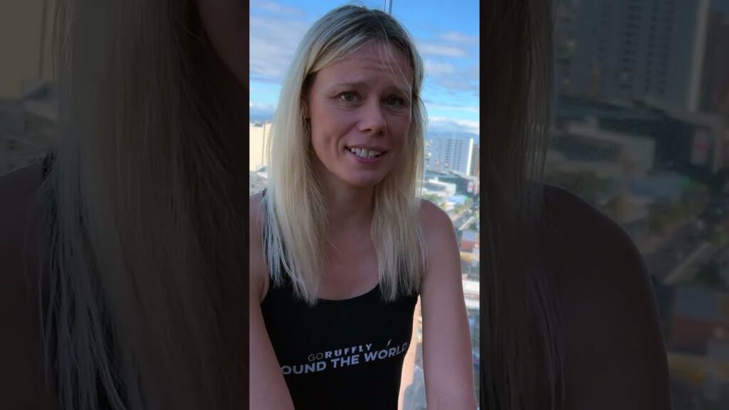 A woman with long blonde hair stands on a balcony overlooking a cityscape. She wears a black tank top with white text. Buildings and a blue sky are visible in the background.