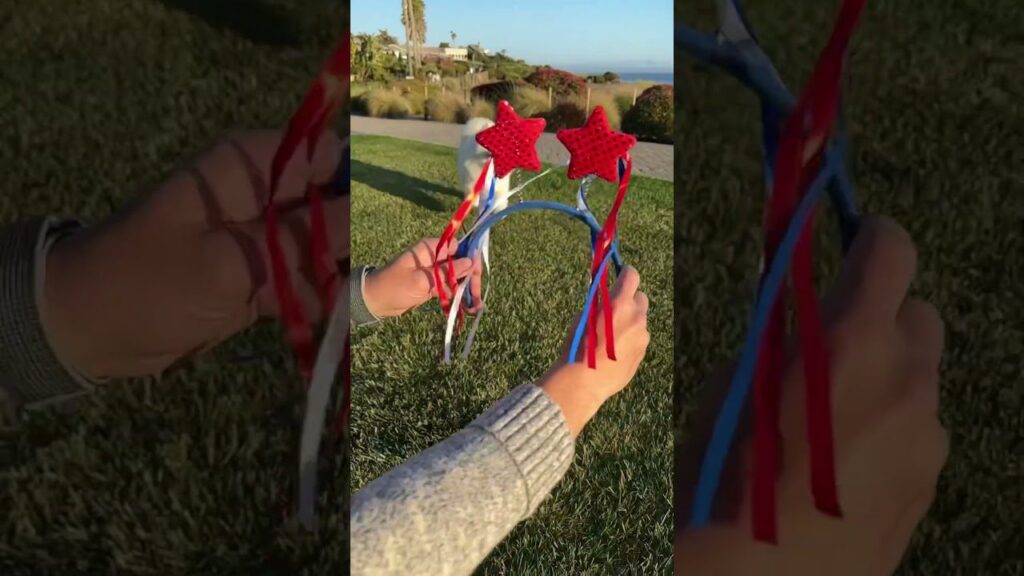 A person holds a pair of headbands adorned with red star decorations and hanging red, white, and blue ribbons. They are standing on a grassy area with a walkway and trees in the background.