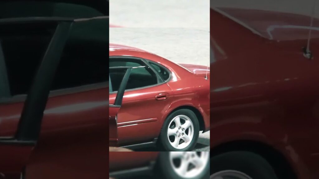 A red sedan with an open rear door is parked on a light gray concrete surface. The car's shiny exterior and silver alloy wheels are visible. The background is blurred, focusing attention on the car's side.