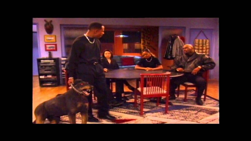 A group of four people sit around a table in a dimly lit room, engaged in conversation. A man stands with a large dog on a leash. The room is decorated with framed pictures and warm-toned furniture.