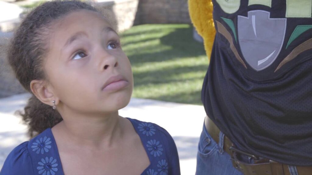 A young girl with curly hair looks up with a curious expression. She is wearing a blue dress with floral designs. Beside her is someone in a graphic t-shirt, partially visible. They are outside on a sunny day.