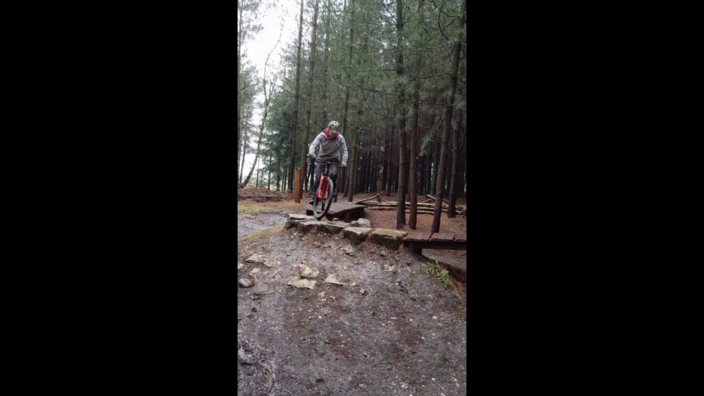 A person wearing a gray hoodie and helmet rides a unicycle down a rocky path in a forest. Tall trees surround the trail, and the ground is covered in dirt and fallen leaves. The atmosphere is slightly overcast.