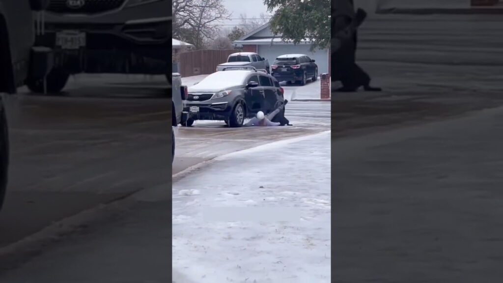 A person crouches down on an icy driveway next to a black car, which appears stuck. Snow covers the ground, and several cars are parked nearby. It is a cold, wintery day.